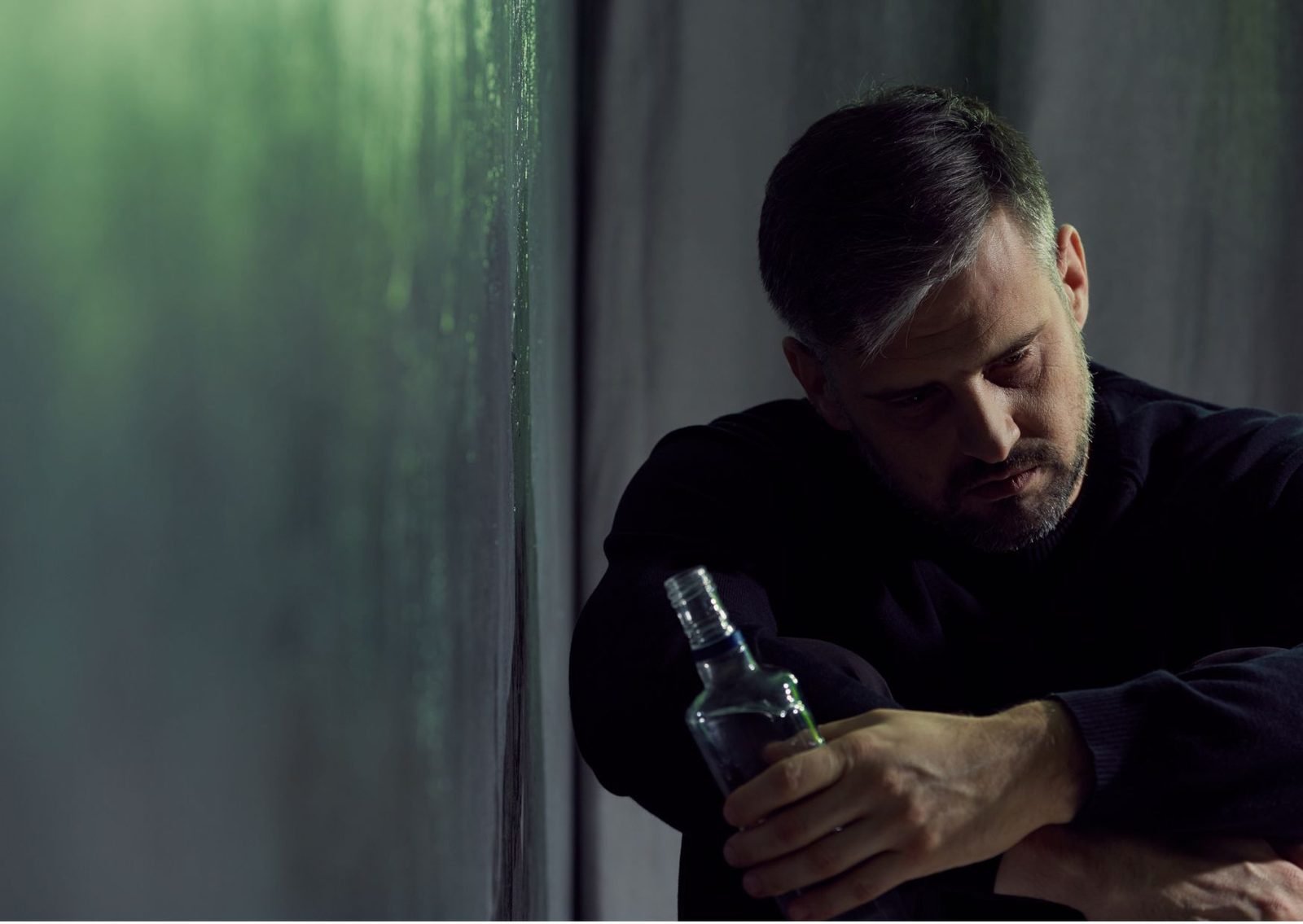 A man sitting alone against a wall, holding a bottle