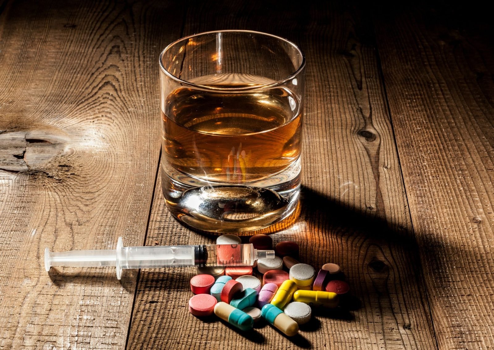 A glass of alcohol beside a syringe and loose pills on a wooden table